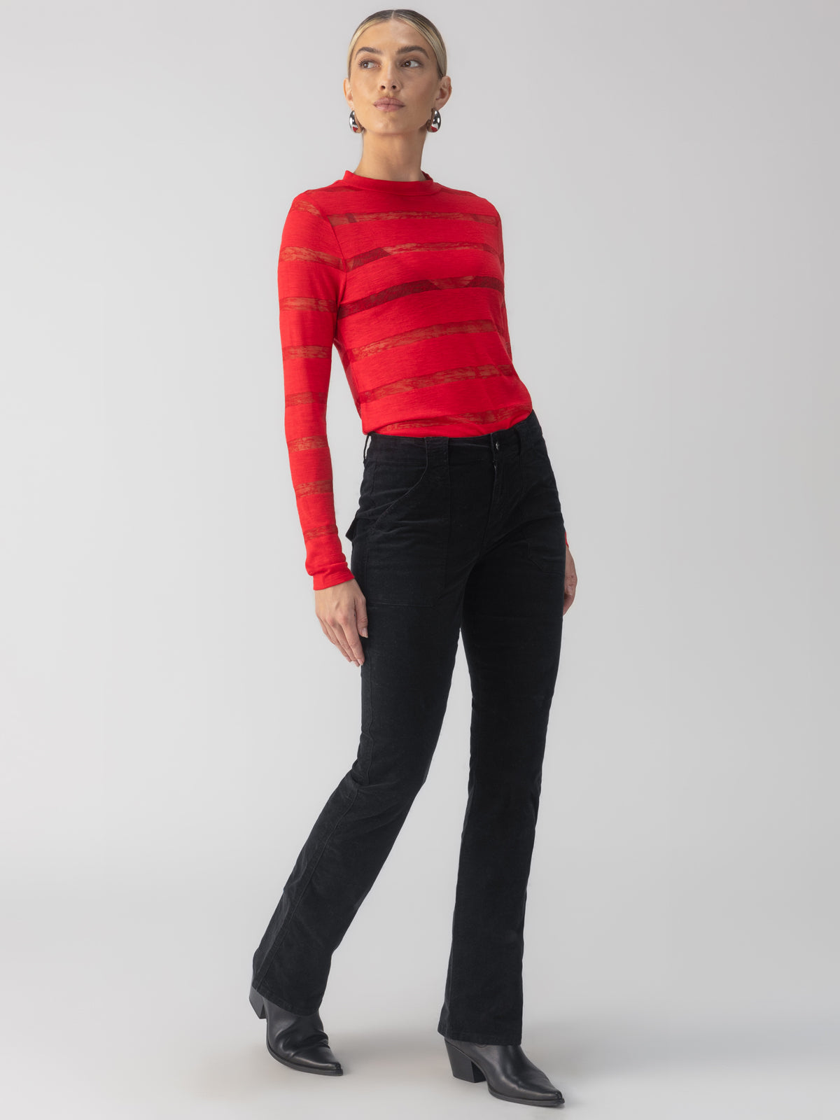 A person wearing the Sanctuary Clothing Semi Sheer Stripe Tee in Mars Red with black pants and heeled boots stands against a light gray background. Their hair is styled back, and they are accessorized with hoop earrings.