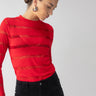 A person wearing the Sanctuary Clothing Semi Sheer Stripe Tee in Mars Red and black pants is looking down. Their hair is pulled back, and they are accessorized with large, round earrings against a plain, neutral background.