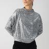 A person wearing the Sanctuary Clothing Sequin Funnel Neck Top in Heather Grey and black pants stands against a plain white background, looking to the side with a subtle smile. Their hair is slicked back, and they have small hoop earrings.