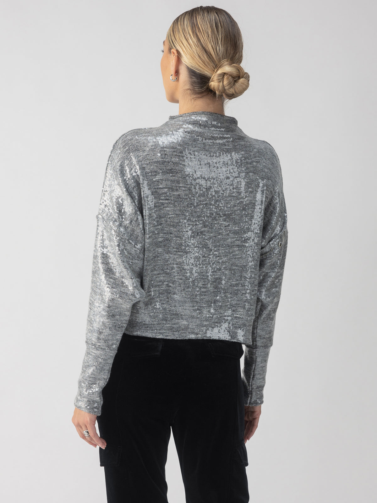 A person with their hair in a bun is wearing the Sanctuary Clothing Sequin Funnel Neck Top in Heather Grey with black pants, viewed from behind against a plain background.