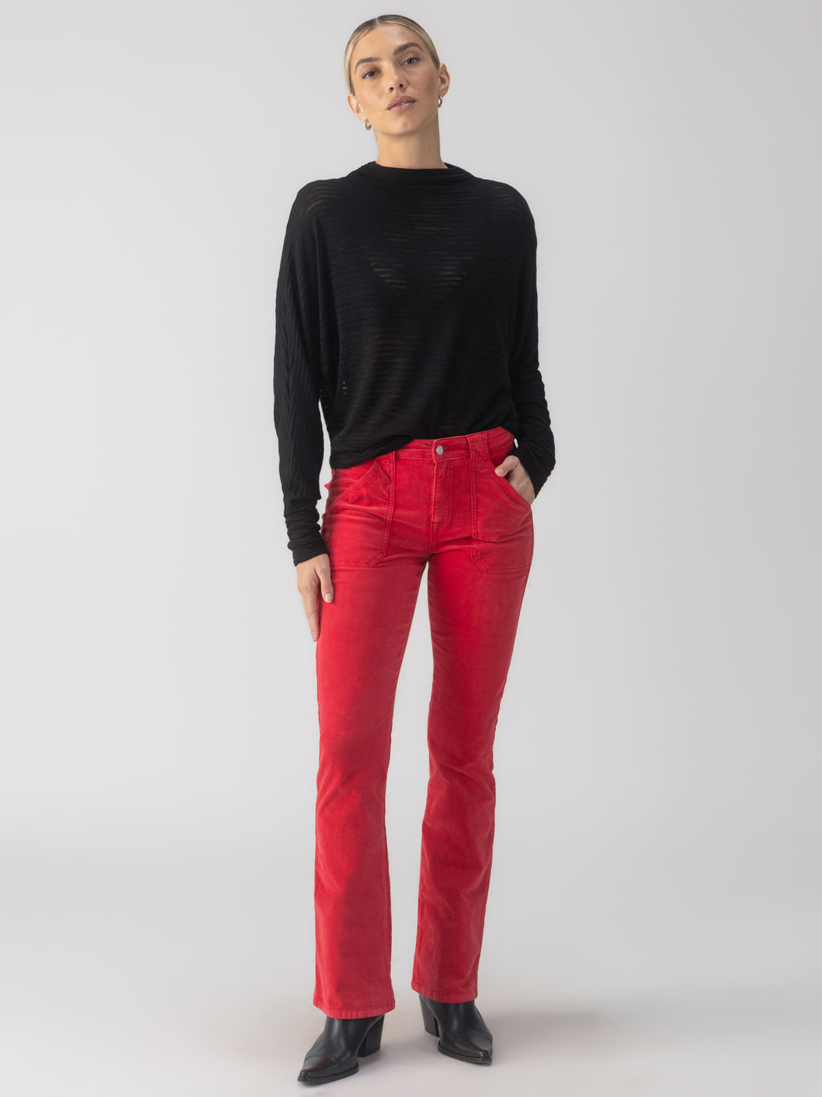 A person with short hair stands against a plain background wearing the Sheer Stripe Funnel Neck Top in black by Sanctuary Clothing, bright red pants, and black heeled boots. They have one hand in a pocket and their head slightly tilted.