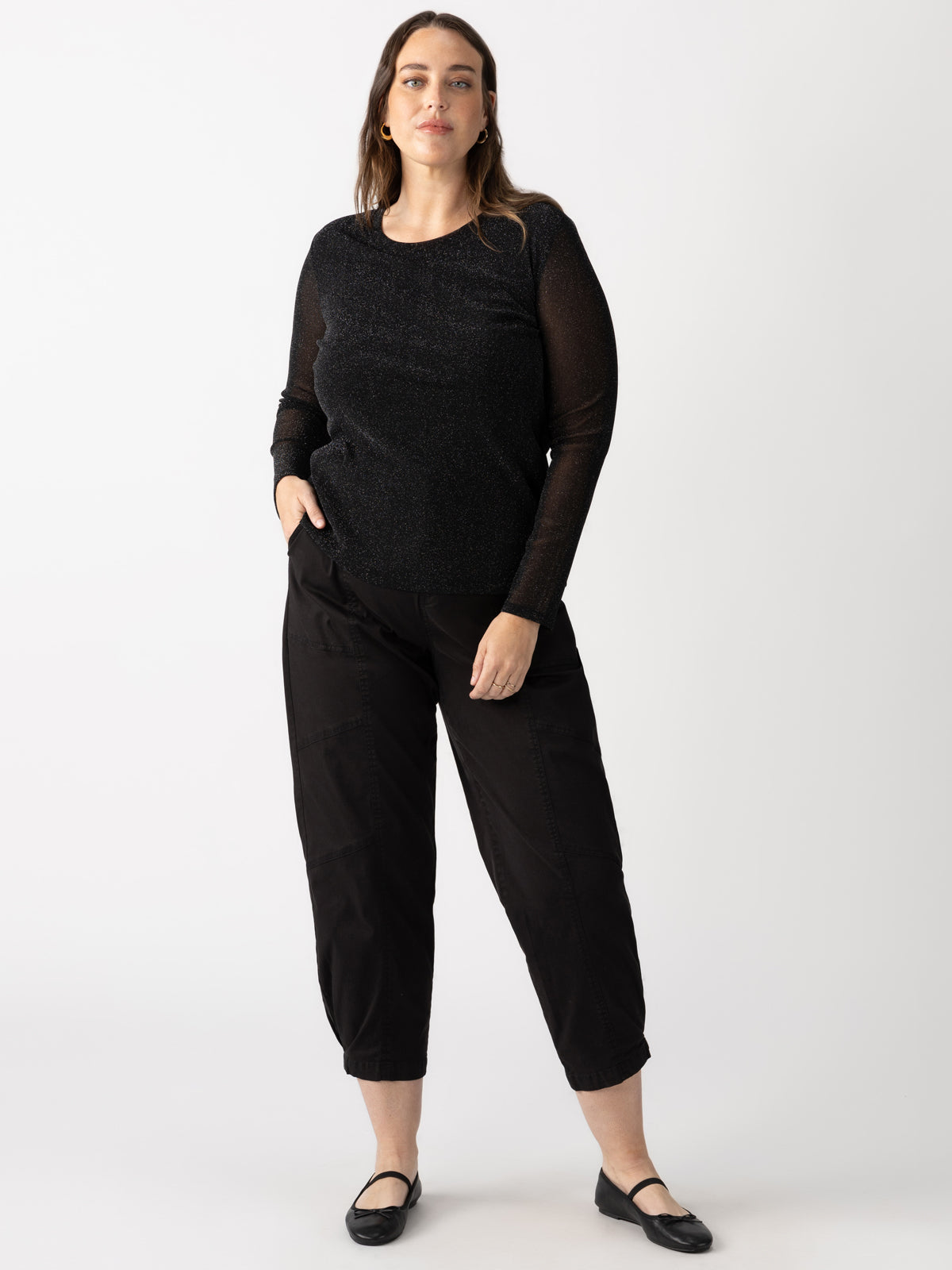 A person dressed in a black long-sleeve top from the Sanctuary Clothing sparkle mesh perfect tee plus size collection and black pants stands against a plain white background. They have one hand in their pocket and are wearing black flats.