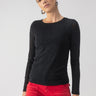 A person dressed in a black "sparkle mesh perfect tee" from Sanctuary Clothing and vibrant red pants stands against a plain gray backdrop. They have short, slicked-back hair, are wearing hoop earrings, and display a neutral, relaxed expression.