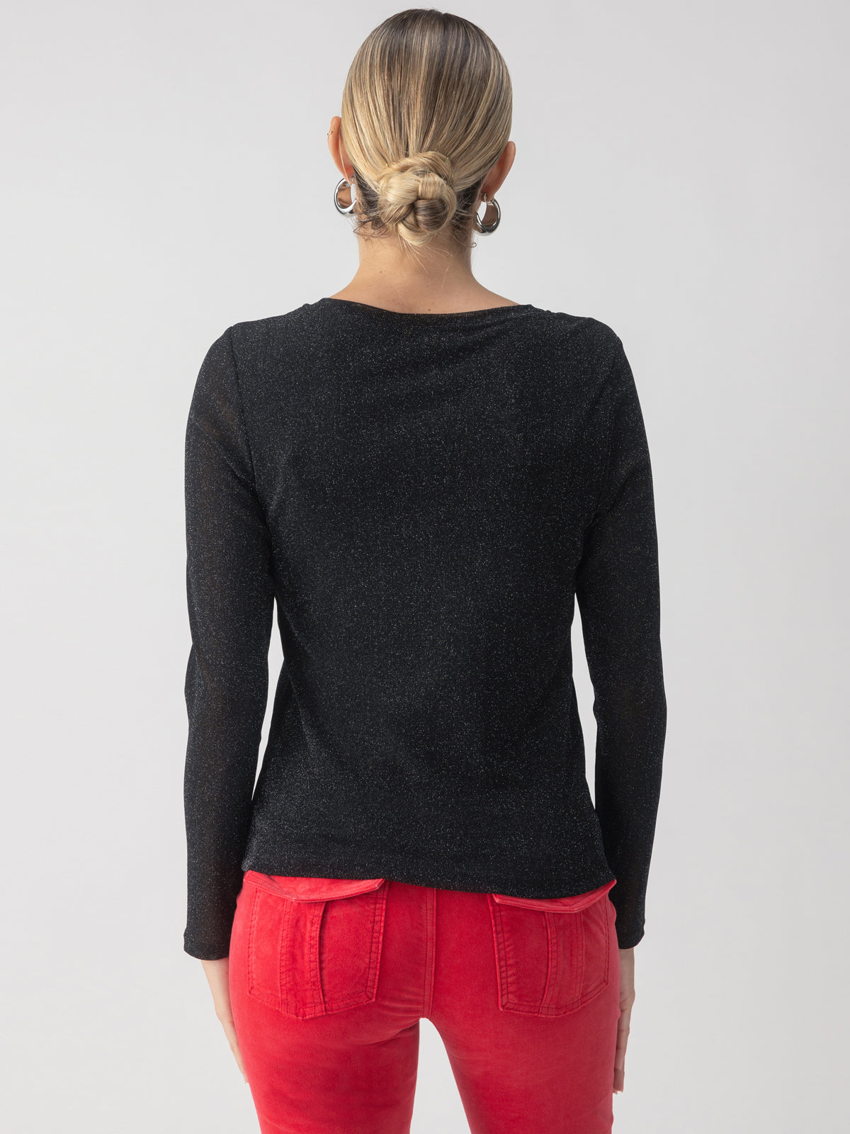 A person with their back to the camera is wearing the Sparkle Mesh Perfect Tee in black by Sanctuary Clothing, along with bright red pants. Their hair is styled in a low bun, and they're standing against a plain gray background.