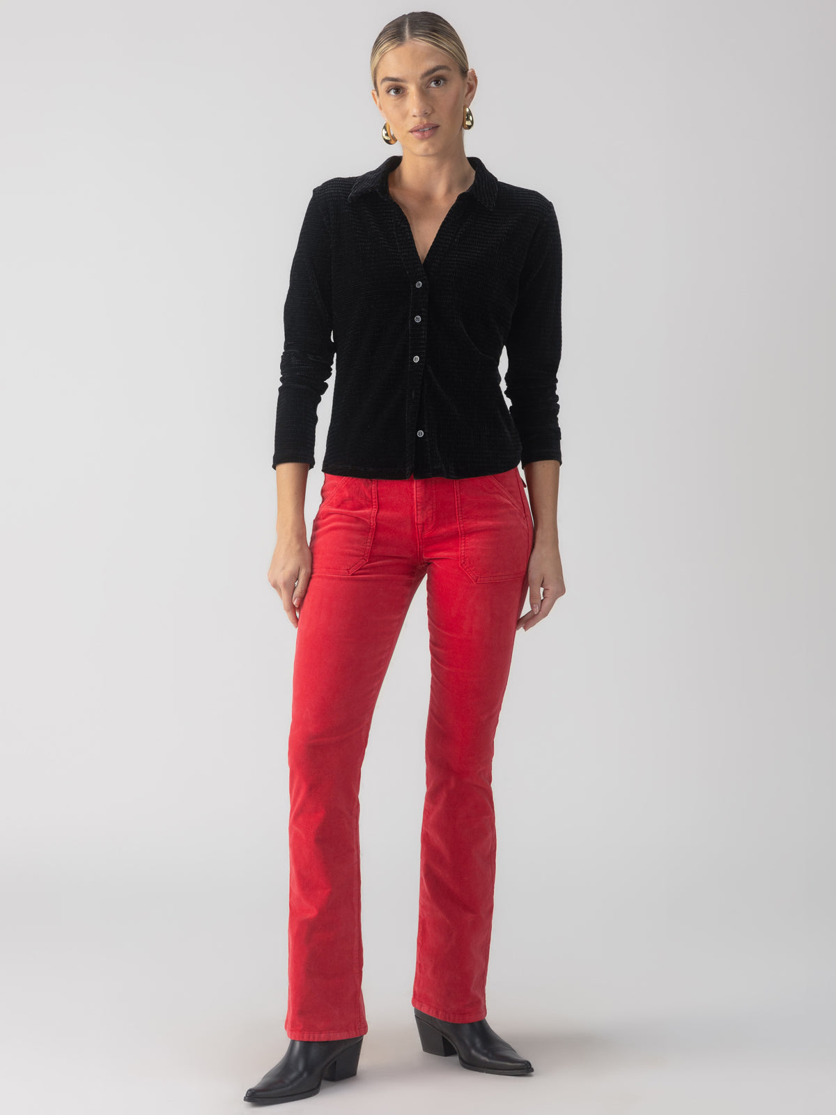 A person with tied-back hair stands against a plain background, wearing a textured velvet button-up top from Sanctuary Clothing, red corduroy pants, and black heeled boots. Their hands are relaxed by their sides as they face forward.