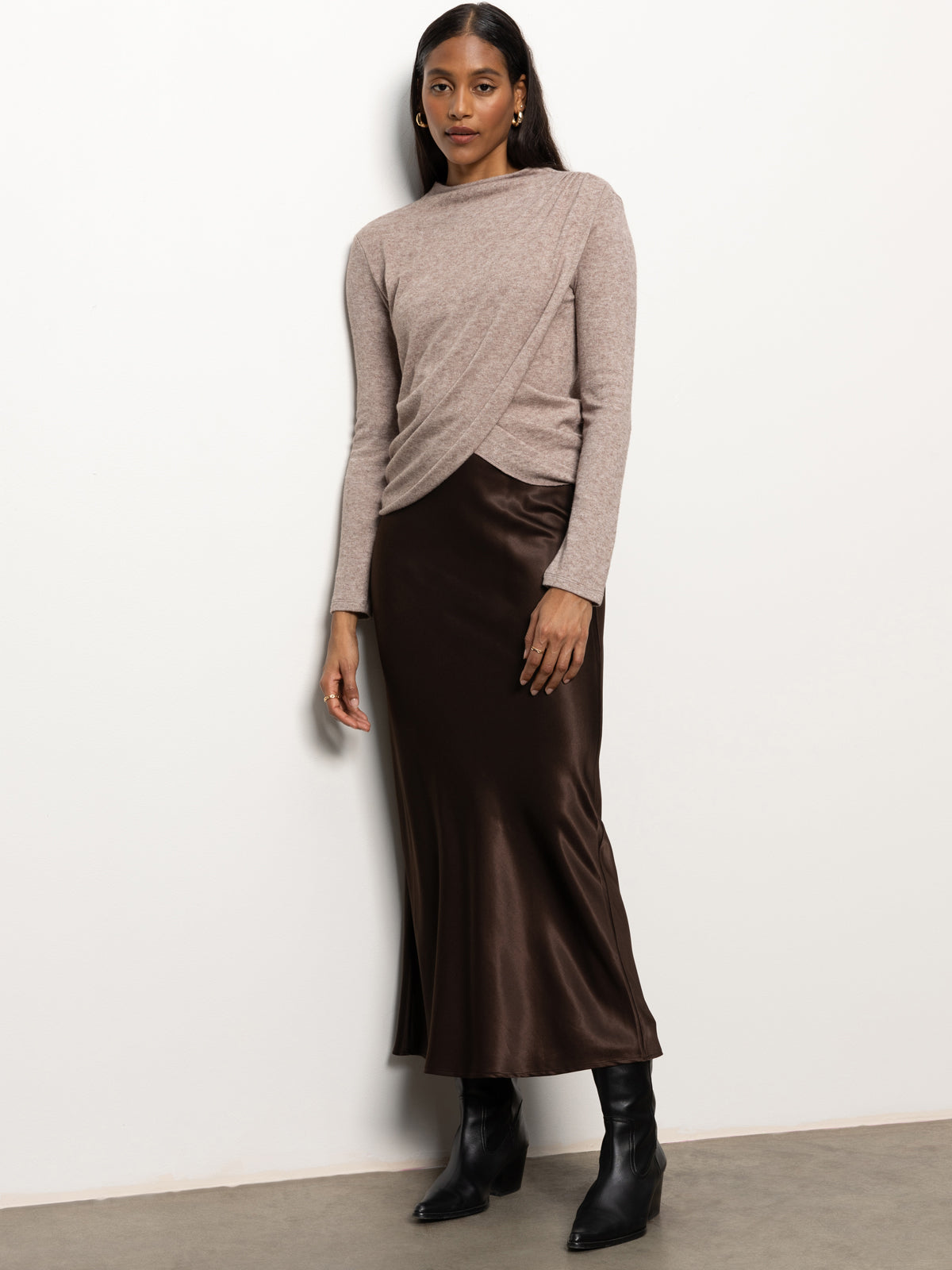 A woman in a Sanctuary Clothing long-sleeve mock draped top in heather cocoa and a dark brown satin skirt stands against a plain white background. She is also wearing black ankle boots and has long, dark hair.