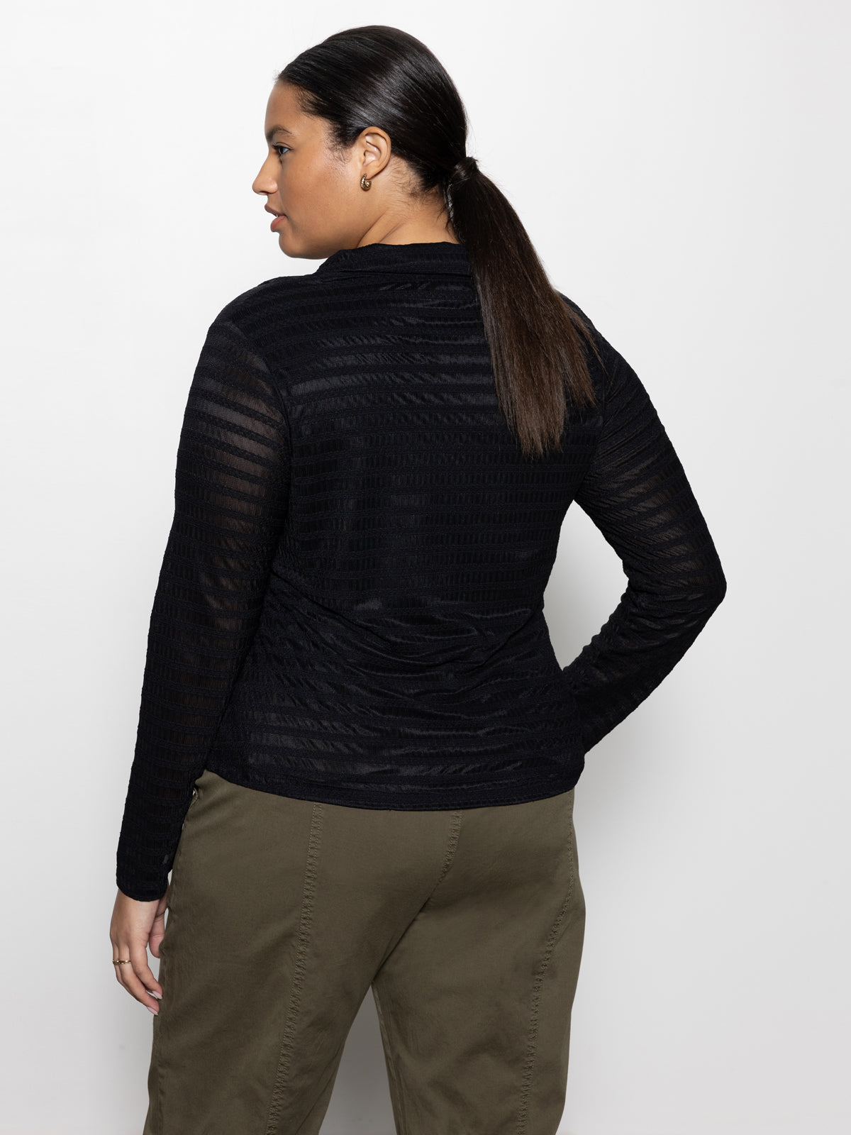 A person stands against a white background, facing away, wearing Sanctuary Clothing's Mesh Lace Shirt Black from the Inclusive Collection. Their hair is tied back in a ponytail, and they pair it with olive green pants.