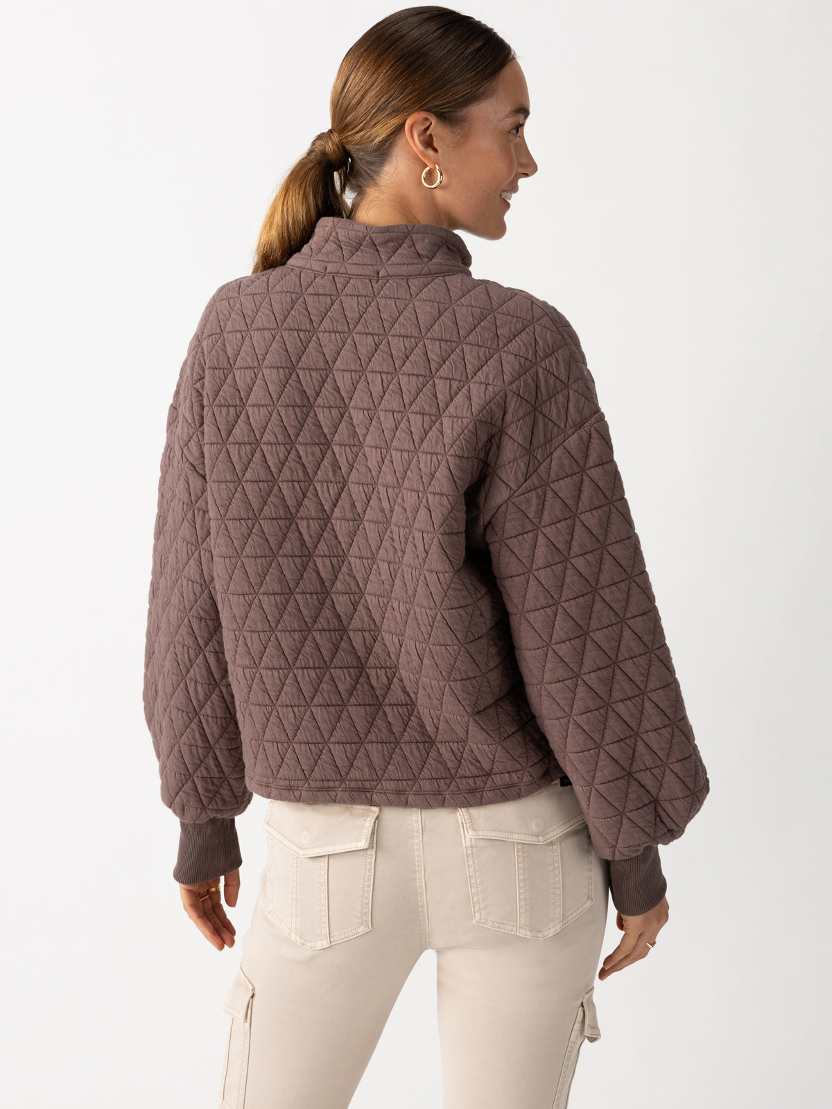 A woman with brown hair in a ponytail is wearing the Quilted Button Up Popover Top Cocoa by Sanctuary Clothing, which features a diamond pattern quilting. She is standing with her back to the camera while wearing light-colored pants against a plain white background.