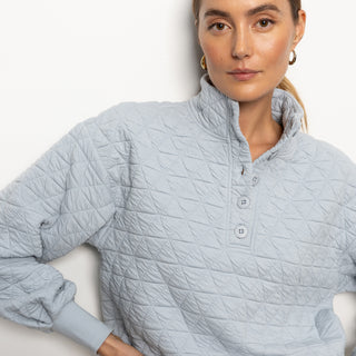 A person with brown hair tied back is wearing the Quilted Button Up Popover Top in Pearl Blue from Sanctuary Clothing. They are standing against a plain white background, looking directly at the camera with a neutral expression.