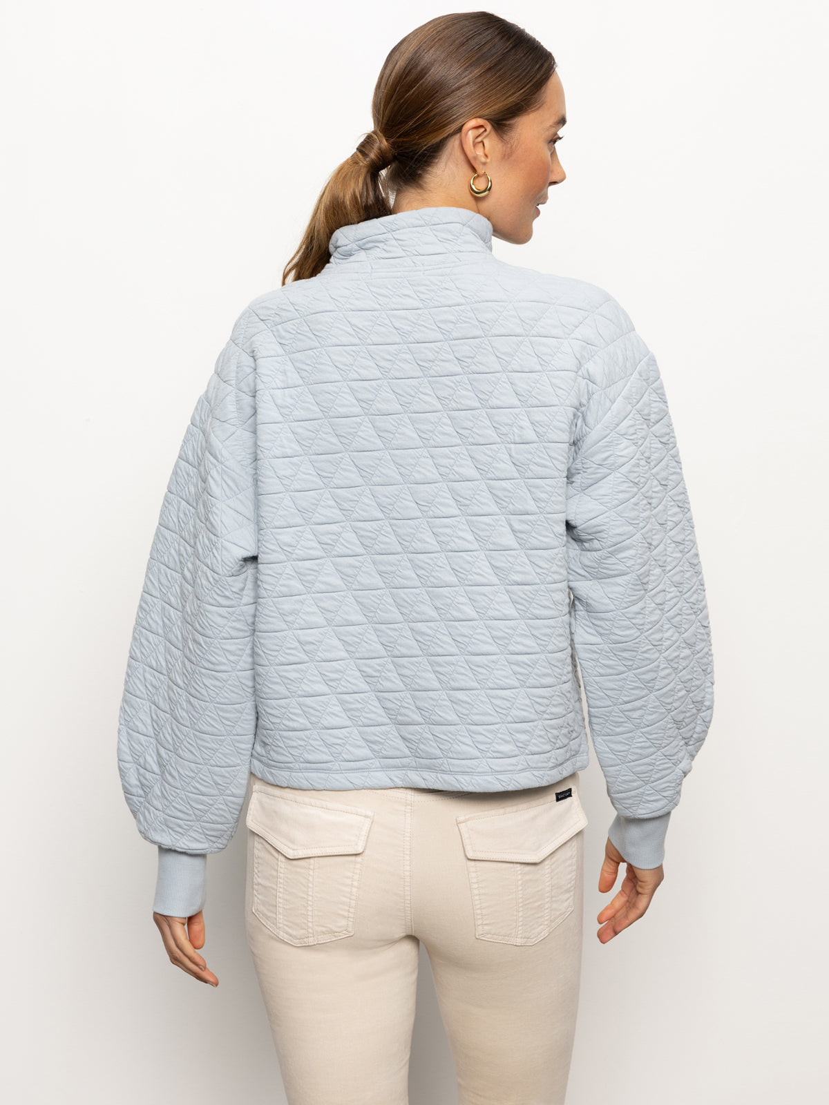 A woman with brown hair in a ponytail is wearing the "Quilted Button Up Popover Top" in Pearl Blue by Sanctuary Clothing and beige pants. She is facing away, against a plain white background.