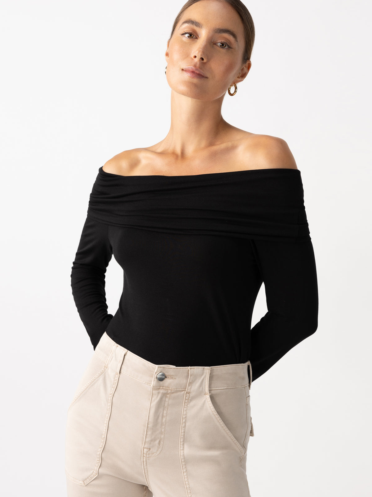 A woman dressed in a Sanctuary Clothing Off The Shoulder Top in black and beige pants, with her hands in her pockets. She's accessorized with hoop earrings and has her hair styled into a bun, posed against a plain background.