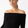 A person with long hair is wearing the Off The Shoulder Top in black from Sanctuary Clothing. They are looking toward the camera with a neutral expression against a plain, light-colored background.