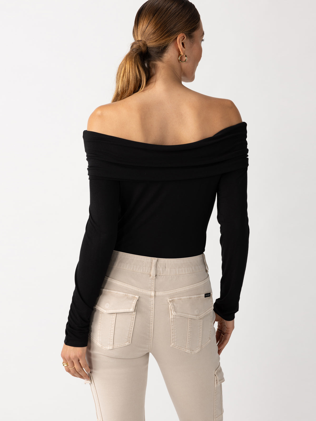 A person is seen from the back wearing the Sanctuary Clothing Off The Shoulder Top in black, paired with beige pants featuring pockets. Their hair is styled in a ponytail, against a plain, light-colored background.