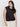 A woman with long dark hair wears Sanctuary Clothing's black knotted muscle tee (extended sizing) and light pants, standing against a plain white background, looking confidently at the camera.