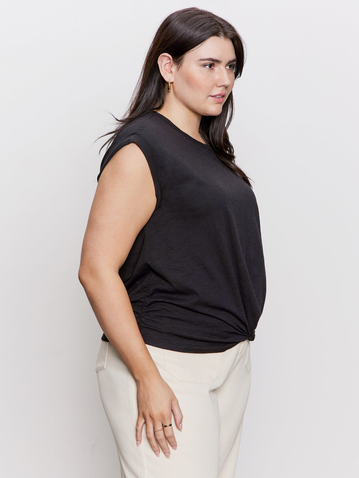A woman with long dark hair wears Sanctuary Clothing's knotted muscle tee in black, suitable for extended sizing, paired with light-colored pants. She stands sideways against a plain white background, looking forward.