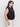 A woman with long dark hair wears Sanctuary Clothing's knotted muscle tee in black, suitable for extended sizing, paired with light-colored pants. She stands sideways against a plain white background, looking forward.