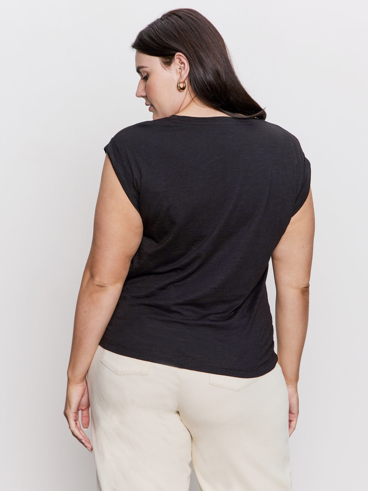 A person with long dark hair is facing away from the camera, wearing Sanctuary Clothing's knotted muscle tee in black from their extended sizing collection and light-colored pants. They have gold hoop earrings and stand against a plain white background.