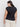 A person with long dark hair is facing away from the camera, wearing Sanctuary Clothing's knotted muscle tee in black from their extended sizing collection and light-colored pants. They have gold hoop earrings and stand against a plain white background.