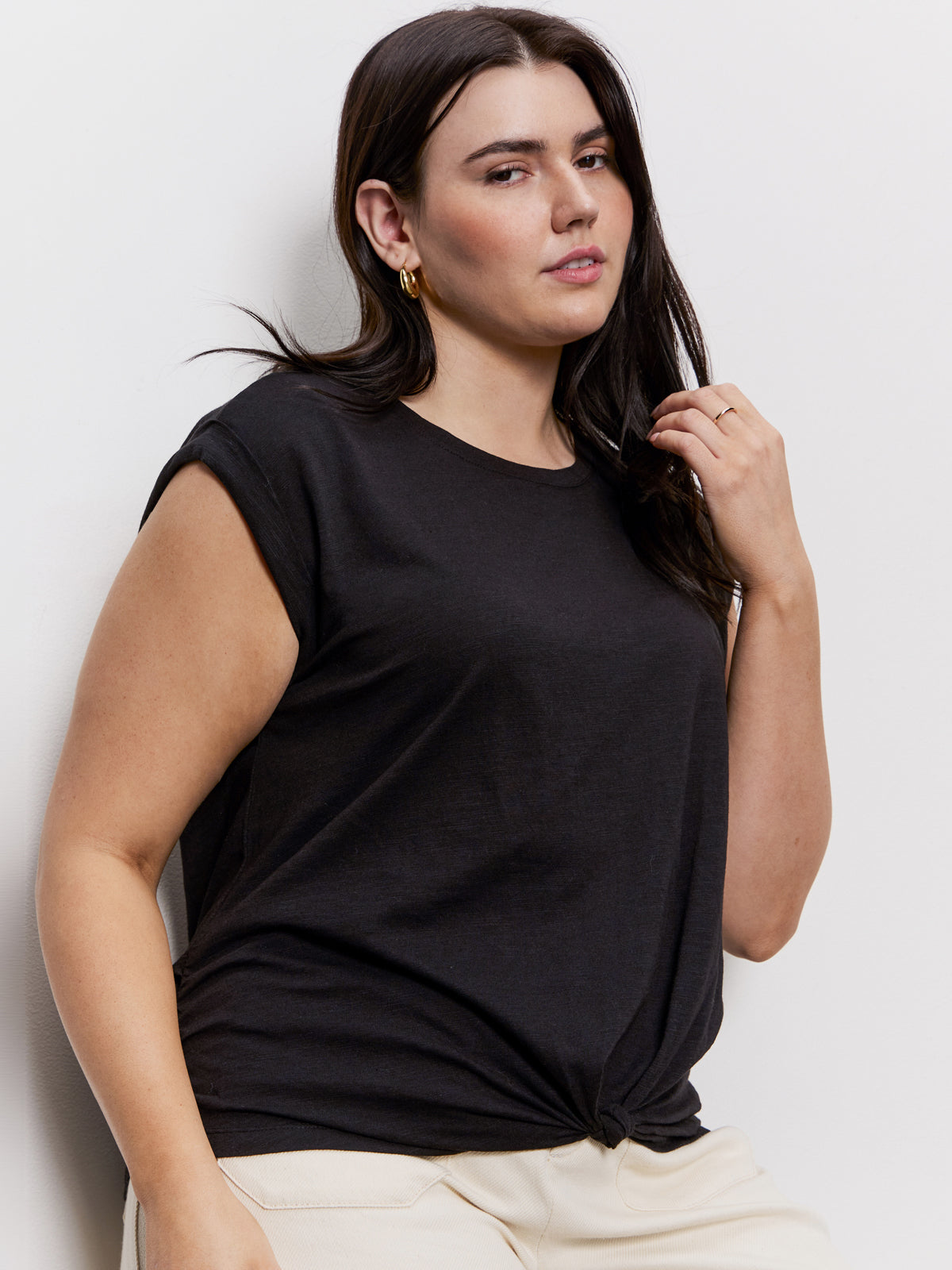 A person with long dark hair wears Sanctuary Clothing's knotted muscle tee in black and cream pants, accessorized with gold hoop earrings. They are gently touching their neck against a plain, light-colored wall backdrop.