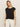 Someone with long hair wears a black knotted muscle tee by Sanctuary Clothing and white pants, standing against an off-white background, looking neutrally at the camera.
