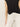 A person wearing Sanctuary Clothing's knotted muscle tee in black with white pants is shown from the shoulders down against a plain light background. Only one arm is visible, with the hand relaxed by their side.