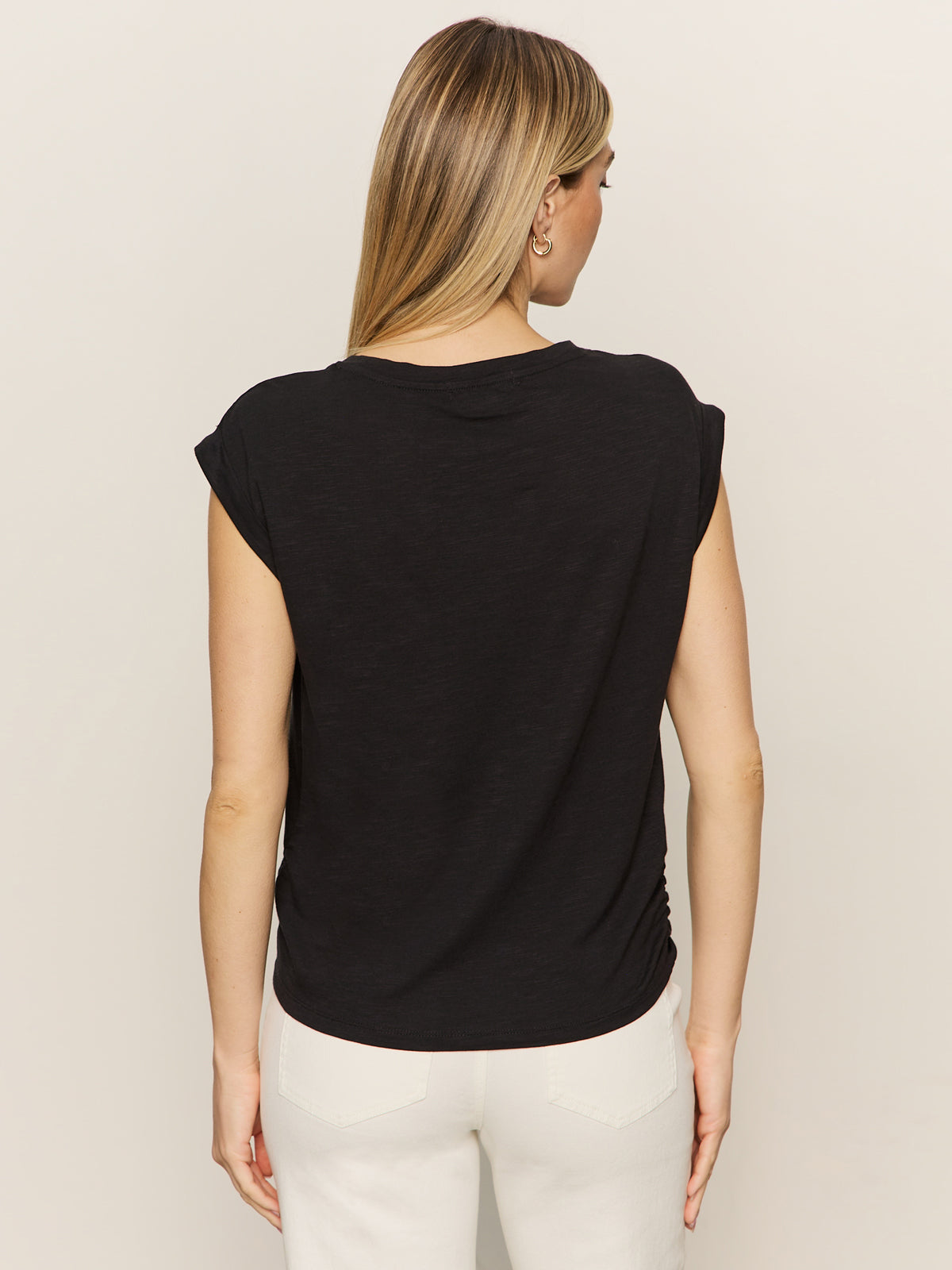 A blonde woman stands facing away, wearing the Sanctuary Clothing knotted muscle tee in black and white pants against a plain, light-colored background.