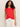 A woman with long curly hair wears the Sanctuary Clothing knotted muscle tee in cayenne and loose white pants, standing against a plain light background with her hands in her pockets.