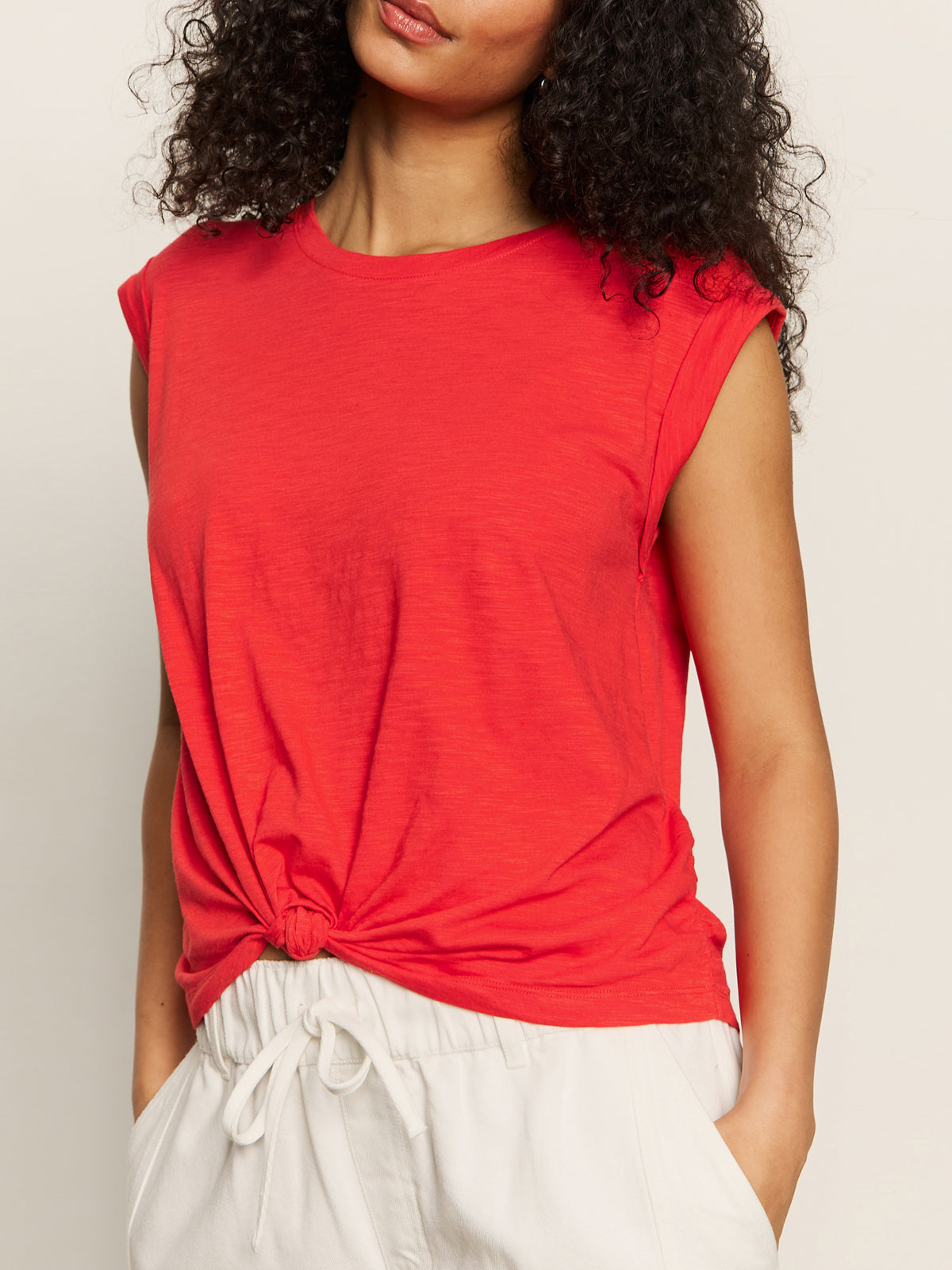 A woman with curly hair wears the Sanctuary Clothing knotted muscle tee in cayenne and white drawstring pants, standing against a plain background with her face partially out of frame.
