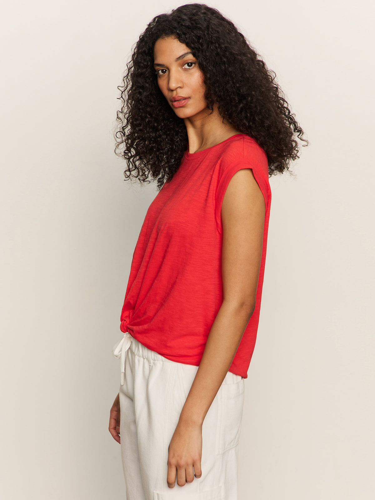 Woman with long curly hair models the Sanctuary Clothing knotted muscle tee in cayenne, styled sleeveless and paired with white pants, standing sideways and looking at the camera against a light background.