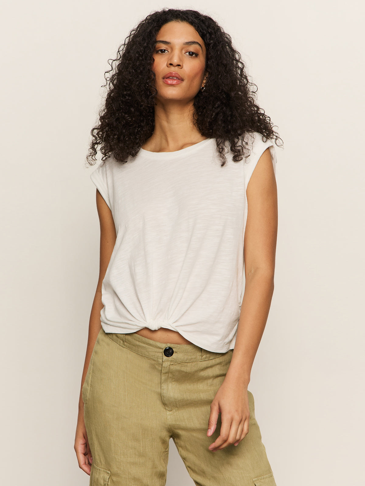 A person with long curly hair wears Sanctuary Clothing's knotted muscle tee in white paired with beige cargo pants, confidently posing against a plain background.