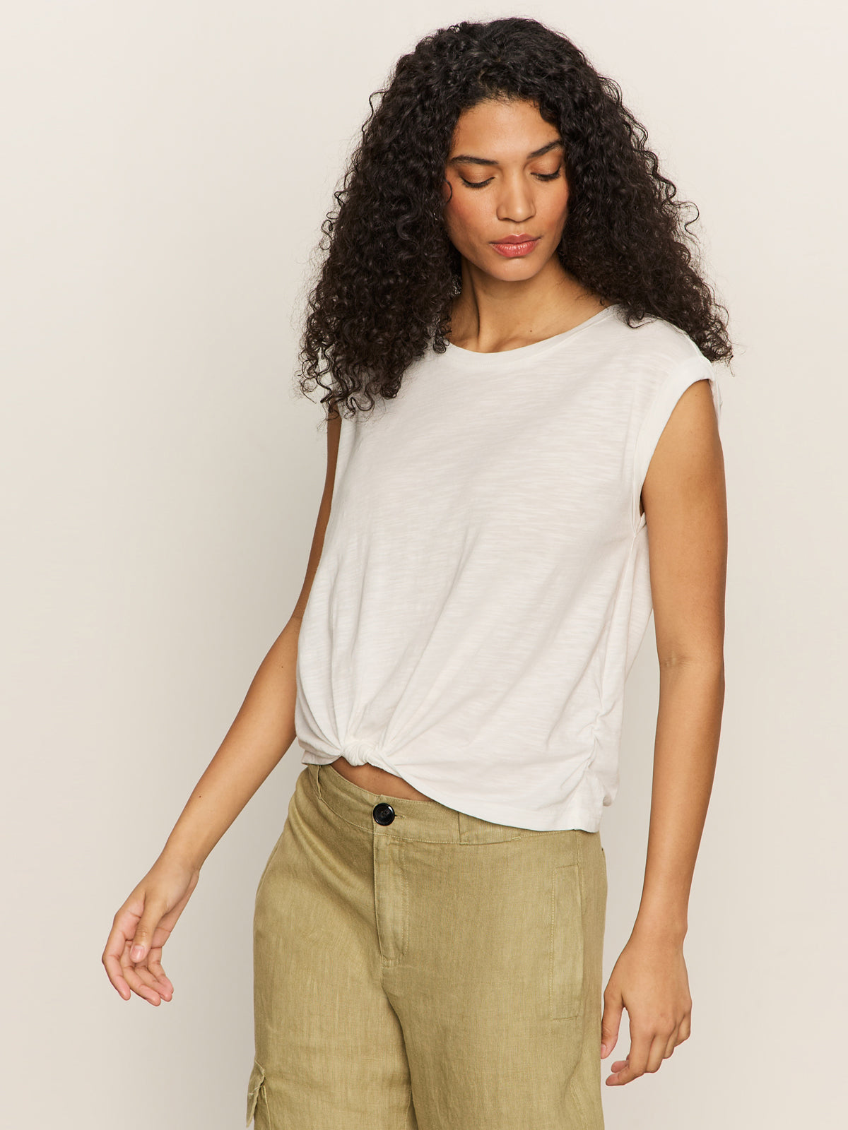 A woman with long curly hair wears Sanctuary Clothing's knotted muscle tee in white and beige pants, looking slightly down and to the side against a neutral background.