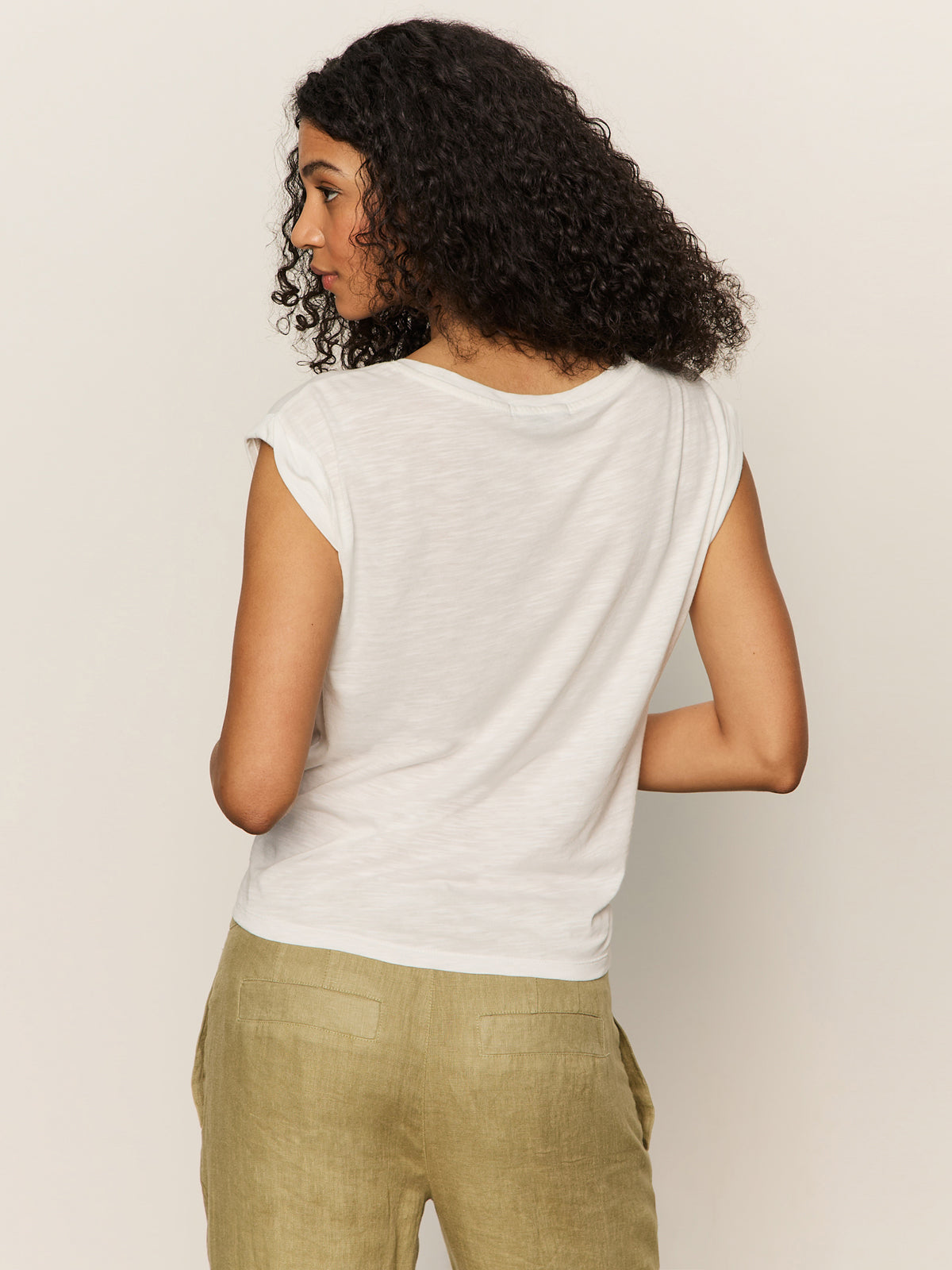 A woman with curly hair stands with her back to the camera in a neutral setting, wearing Sanctuary Clothing's knotted muscle tee in white and light brown pants, gazing over her left shoulder.