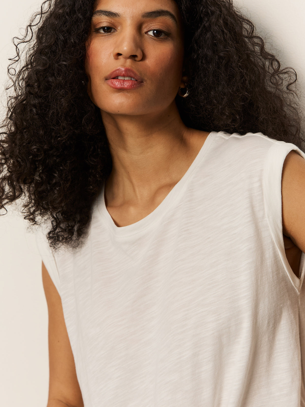 A person with curly black hair is wearing a knotted muscle tee by Sanctuary Clothing. They have a neutral expression while looking slightly to the side against a plain, light-colored background.