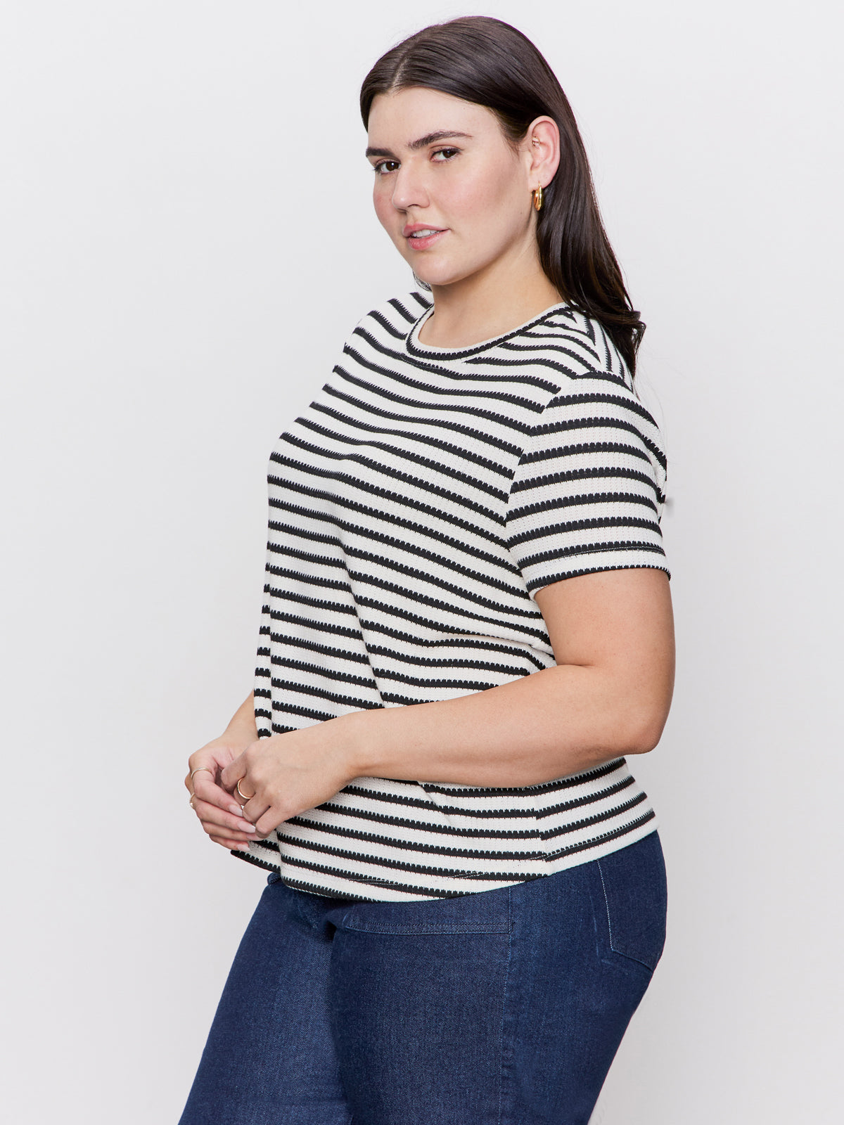 Wearing the Sanctuary Clothing "perfect textured tee macrame black stripe extended sizing" and blue jeans, a person with long dark hair stands sideways, smiling slightly at the camera. Simple jewelry complements their look against a plain, light-colored background.