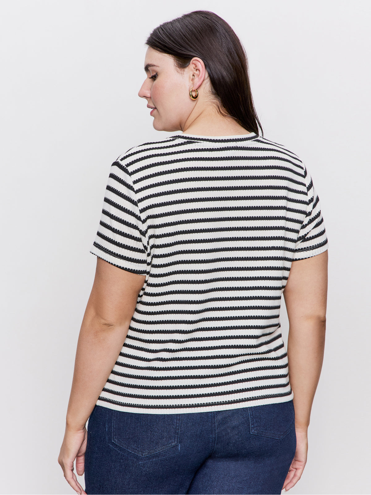 A person with long dark hair faces away, donning Sanctuary Clothing's perfect textured tee in macrame black stripe. They pair it with dark jeans and gold hoop earrings against a plain white background.