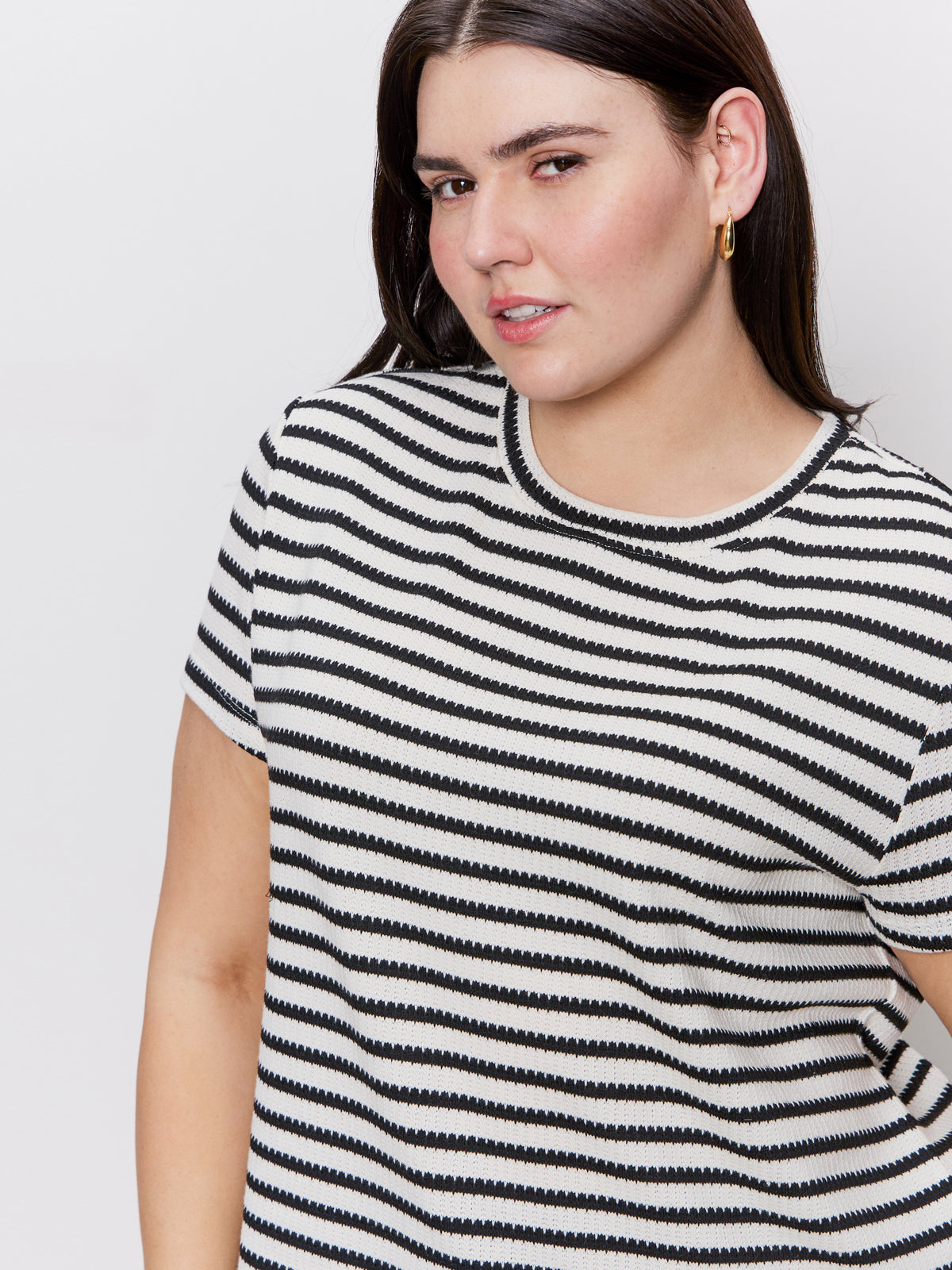 A person with long dark hair, wearing Sanctuary Clothing's "Perfect Textured Tee in Macrame Black Stripe" is looking at the camera with a slight smile against a plain white background, accessorized with gold hoop earrings.