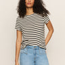 A person with long curly hair, wearing Sanctuary Clothing's "perfect textured tee macrame black stripe" and light blue jeans, stands against a plain background with a relaxed posture and one hand on their hip.