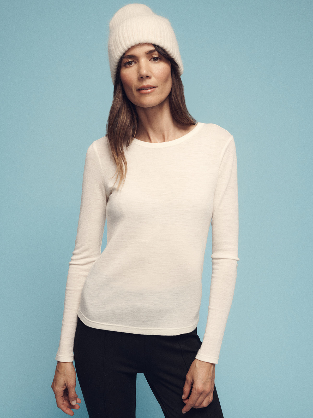 A person wearing the Sanctuary Clothing long sleeve wool perfect tee in chalk and a knitted beanie stands against a light blue background, looking at the camera with a relaxed expression.