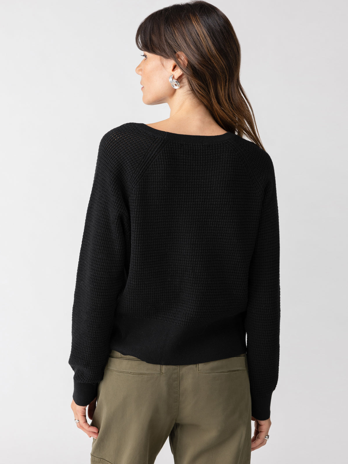 A person with long brown hair is shown from the back, wearing a black Everyday Openwork Sweater by Sanctuary Clothing and olive-green pants. The background is plain and light-colored.