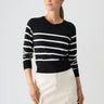 A person with long hair is gazing to the side while wearing the Sanctuary Clothing All Day Long Sweater in Black Chalk Stripe and a light-colored skirt with pockets. The individual, adorned with earrings and a necklace, stands against a plain white background.