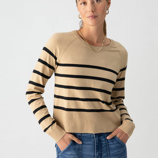 A woman stands against a plain background, wearing Sanctuary Clothing's All Day Long Sweater in True Khaki Black Stripe paired with blue jeans. She has her hands in her pockets, her hair pulled back, and wears hoop earrings and a chain necklace. Her expression is neutral.