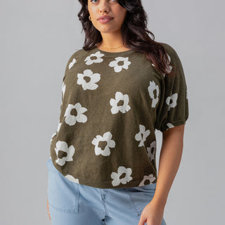 A woman stands confidently against a gray backdrop wearing the Sunny Days Sweater Burnt Olive Pop from Sanctuary Clothing's Inclusive Collection, featuring a relaxed fit and a white floral pattern. She pairs it with light blue pants, has long wavy hair, and accessorizes with small hoop earrings. Her hands are in her pockets.