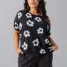 A woman with long, dark hair models the Sunny Days Sweater Flower Pop from Sanctuary Clothing's Inclusive Collection, paired with dark cargo pants and standing against a plain gray background, her hands tucked casually in her pockets.