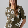 A woman with long brown hair is wearing the Sunny Days Sweater Burnt Olive Pop from Sanctuary Clothing, which is a green short-sleeved sweater with white flower patterns, paired with white pants. She is standing against a plain background, smiling slightly with her hands behind her back.