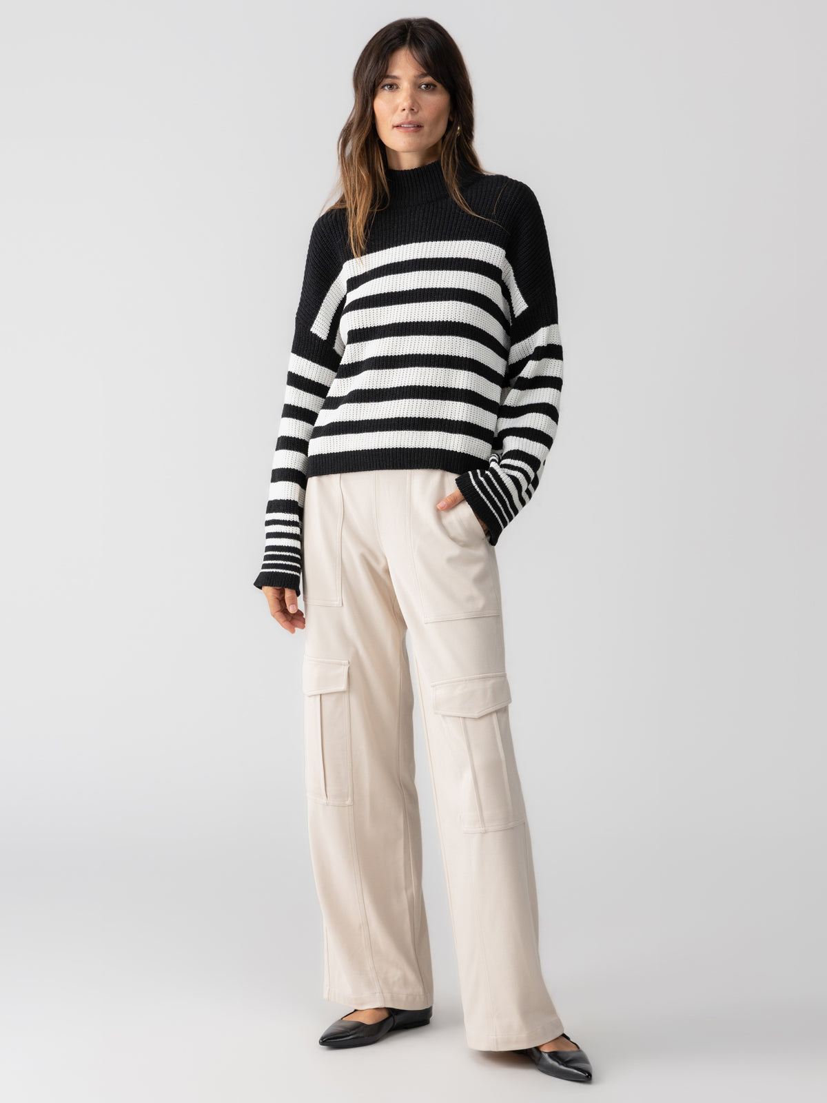 A woman stands against a plain white background, wearing the Stay Cozy Sweater in Black Chalk Stripe by Sanctuary Clothing. She has long brown hair, her hands are in her pockets, and she is paired with light-colored, wide-legged cargo pants and black flats.