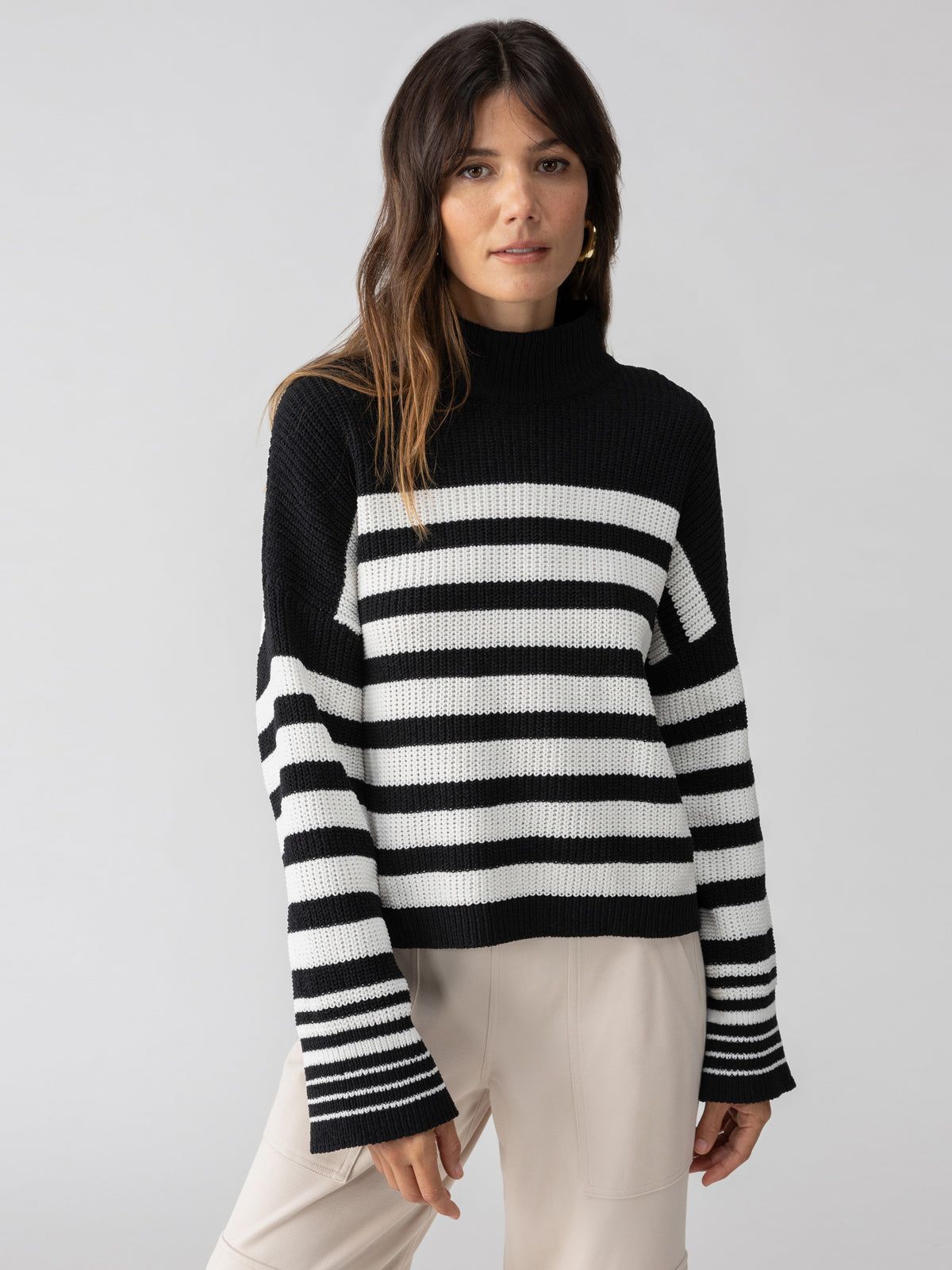 A woman with long brown hair is wearing the Sanctuary Clothing Stay Cozy Sweater in Black Chalk Stripe, featuring a high neckline, paired with light-colored pants. She stands against a plain gray background, looking at the camera with a neutral expression.