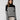 A woman with long brown hair is wearing the Sanctuary Clothing Stay Cozy Sweater in Black Chalk Stripe, featuring a high neckline, paired with light-colored pants. She stands against a plain gray background, looking at the camera with a neutral expression.