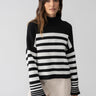 A woman with long brown hair is wearing the Sanctuary Clothing Stay Cozy Sweater in Black Chalk Stripe, featuring a high neckline, paired with light-colored pants. She stands against a plain gray background, looking at the camera with a neutral expression.