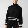 A person with a neat hairstyle is wearing the Stay Cozy Sweater Black Toasted Stripe by Sanctuary Clothing, paired with black pants. The sweater features white stripes near the cuffs and hem. The individual is accessorized with gold hoop earrings and positioned against a plain light-gray background.