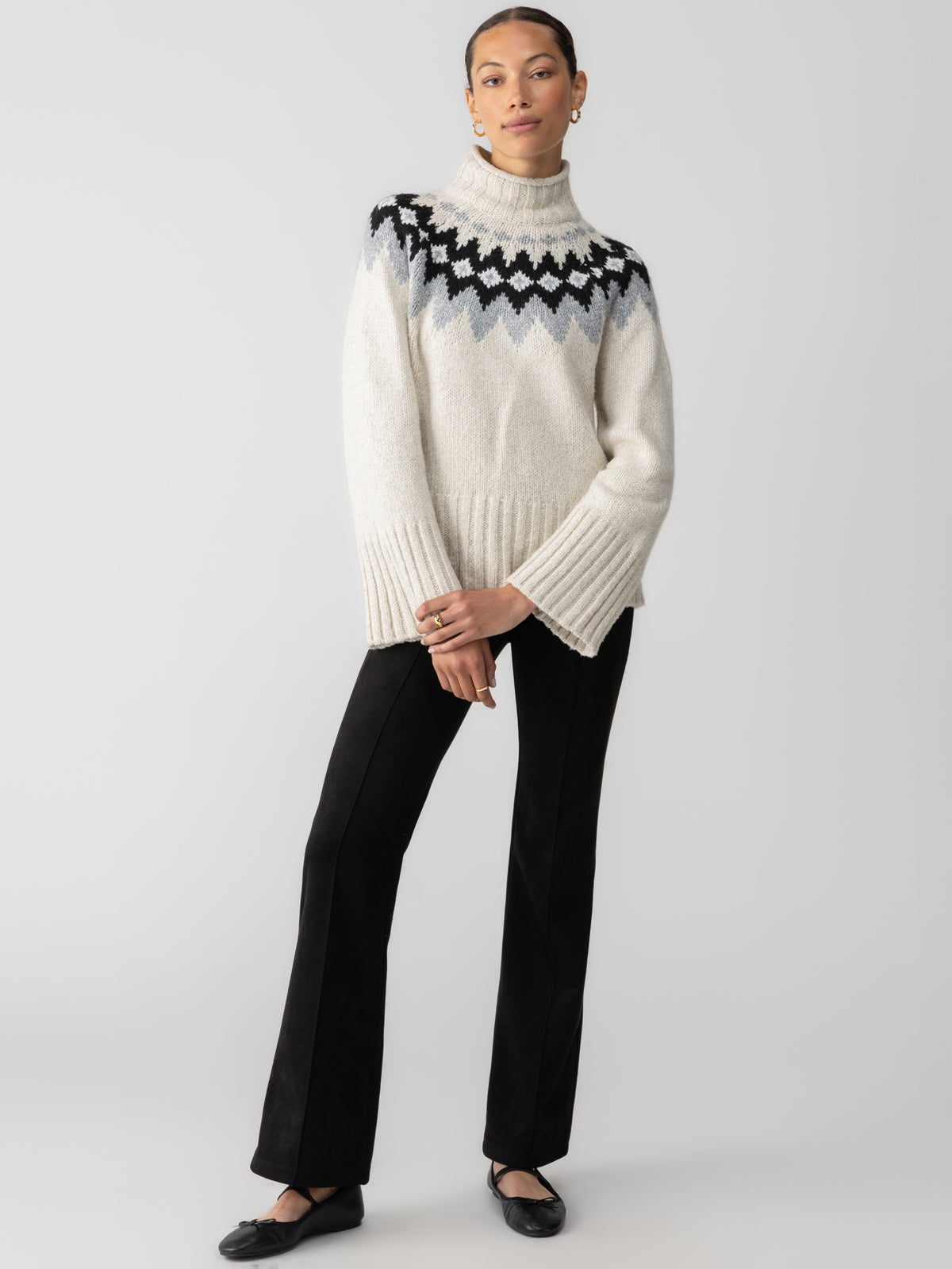 A person is seen posing in the "Tis The Season Fairisle Sweater Toasted Almond Multi" by Sanctuary Clothing. The sweater features a white base with a striking black and gray geometric pattern across the shoulders and chest. They complete their outfit with black pants and black shoes, standing with their hands crossed in front of a plain white background.